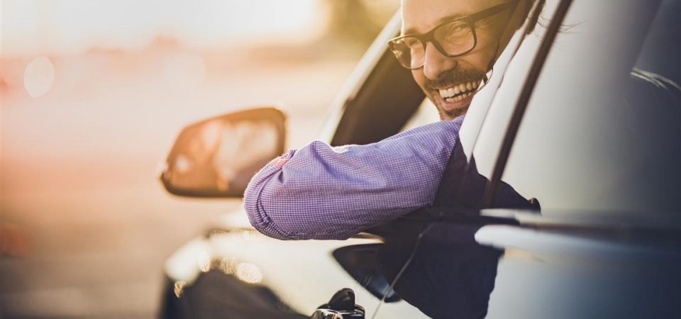 Happy businessman driving a car while going on a trip at sunset and looking at camera.