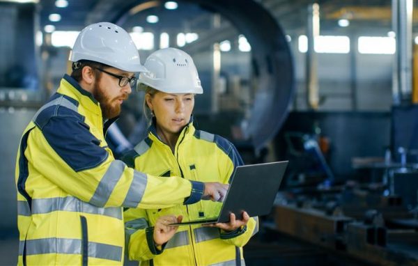 Male and Female Industrial Engineers in Hard Hats Discuss New Project while Using Laptop. They Make Showing Gestures.They Work in a Heavy Industry Manufacturing Factory.